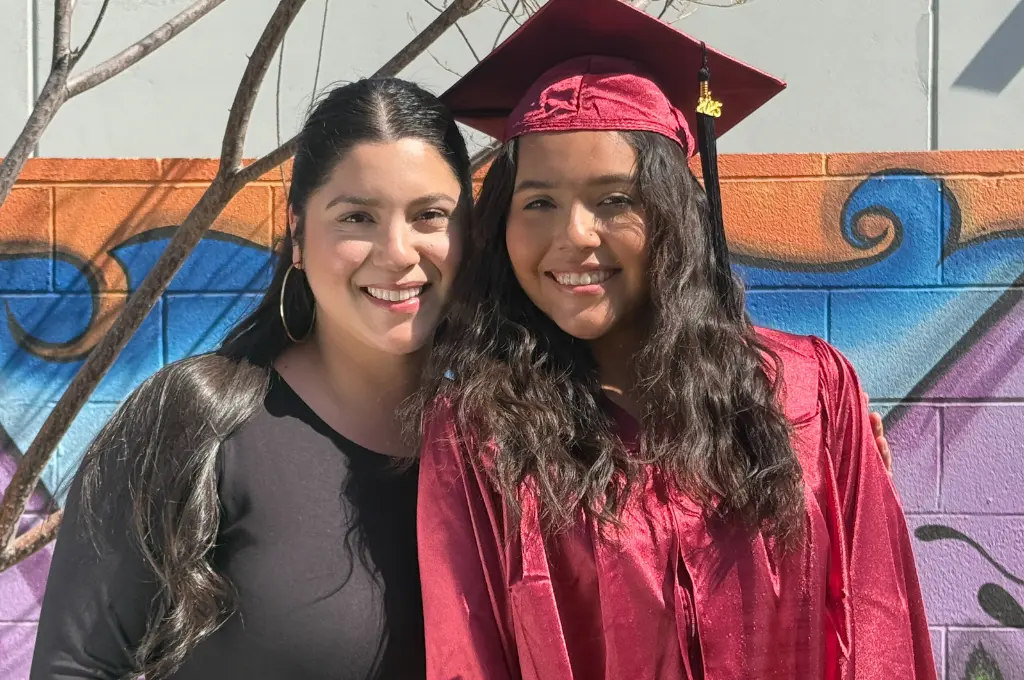 Woman and young woman wearing graduation cap posing together in front of a colorful mural.