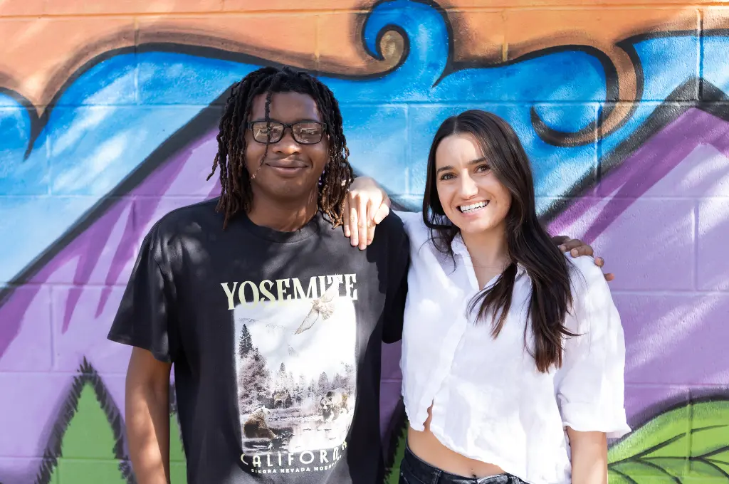 Two young people smiling and posing together against a blue and colorful background.
